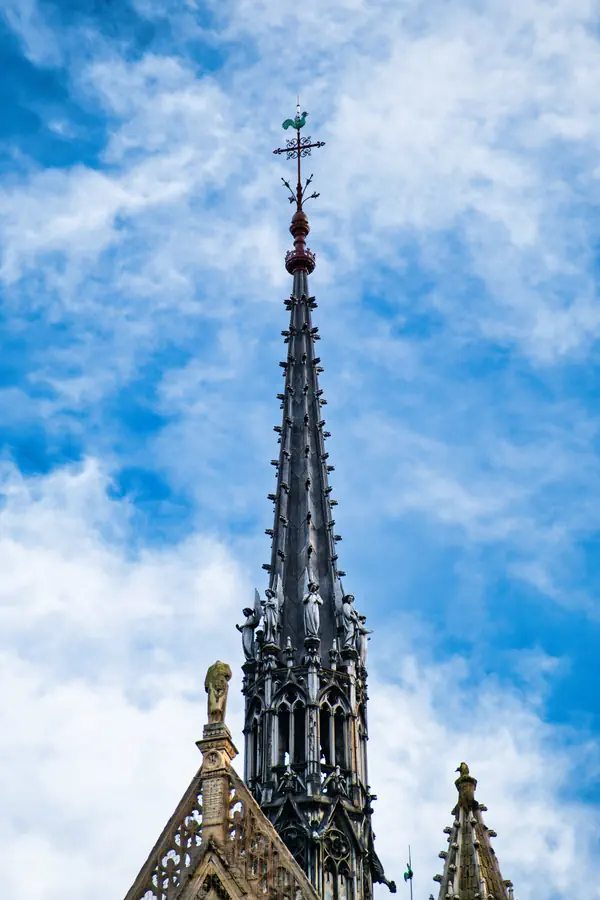 Flèche gothique élancée d'une église, ornée de crochets décoratifs et couronnée d'une croix et d'une girouette verte. La flèche en métal noir est agrémentée de statues et de pinacles, et se détache sur un ciel bleu parsemé de nuages blancs.