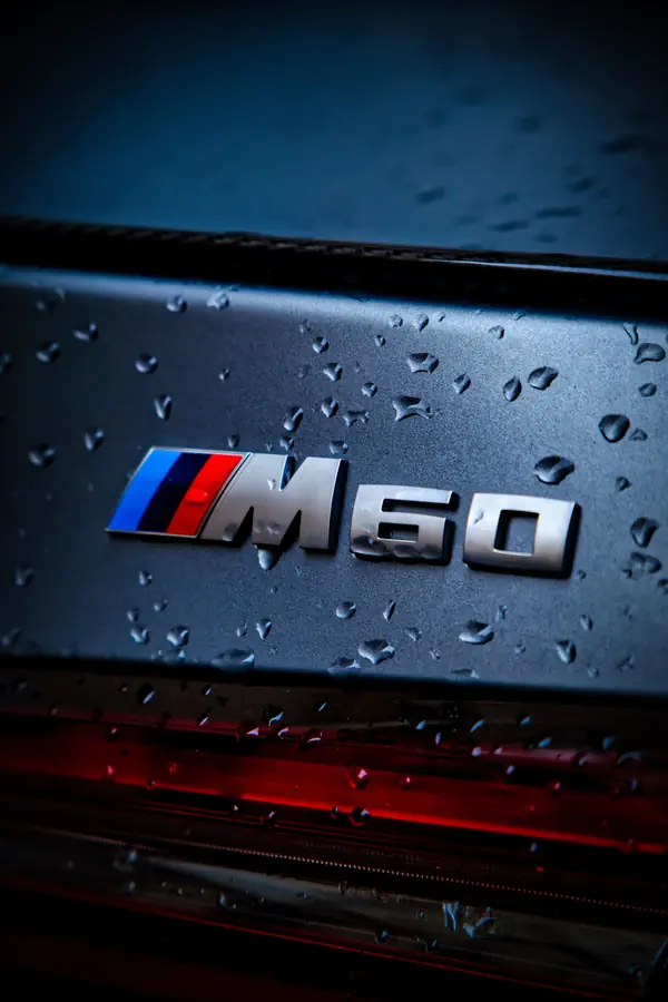 Gros plan sur le badge "M60" d'une BMW, avec le logo M aux couleurs caractéristiques (bleu, violet et rouge) sur une carrosserie gris foncé parsemée de gouttes de pluie.