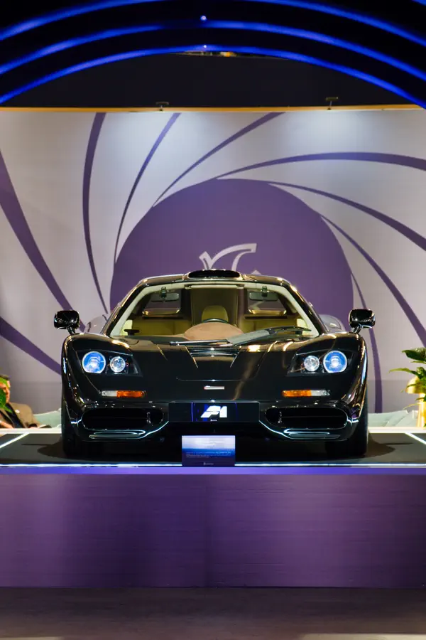 Vue de face d'une McLaren F1 noire sur un podium éclairé, devant un décor graphique circulaire violet.