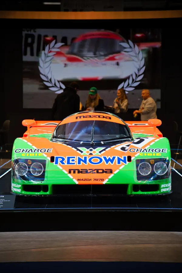 Vue de face d'une Mazda 787B de course arborant sa livrée iconique Renown orange et verte, exposée sur un podium lors d'un événement.