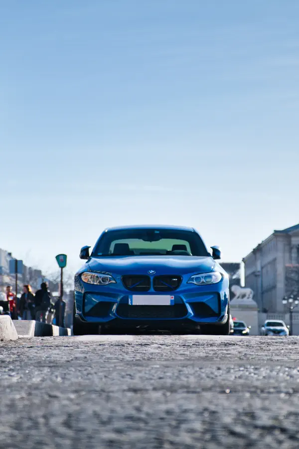 Vue de face d'une BMW M2 bleue centrée sur une route pavée sous un ciel bleu dégagé.