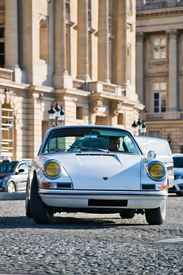 Porsche 911 classique blanche aux phares jaunes roulant sur des pavés devant une façade monumentale en pierre de taille.