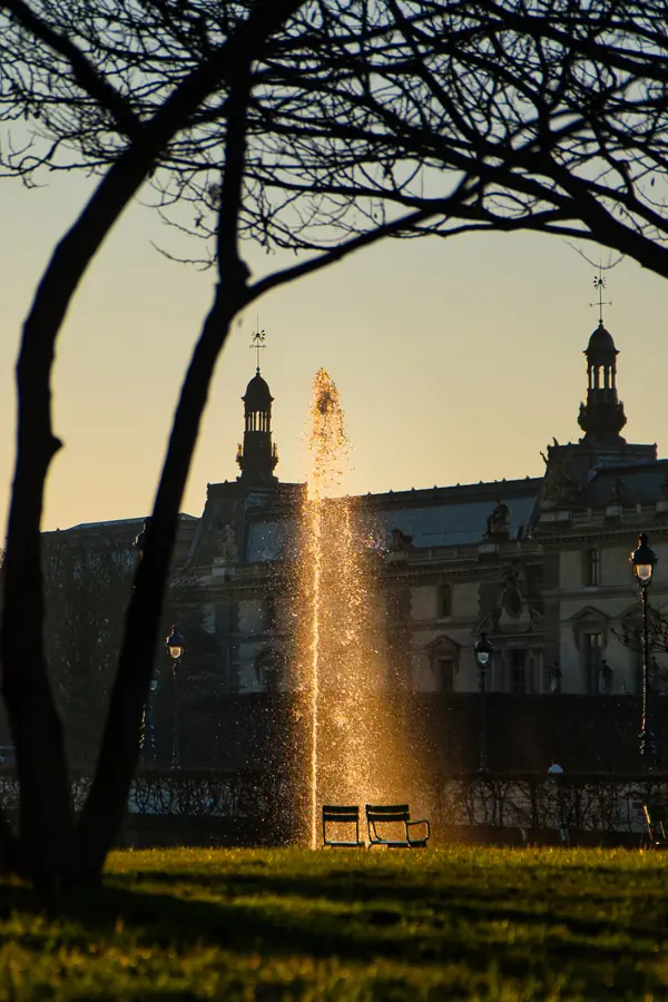 Jet d'eau illuminé par le soleil couchant dans un jardin parisien, créant une colonne dorée devant un bâtiment historique avec ses tourelles caractéristiques.
