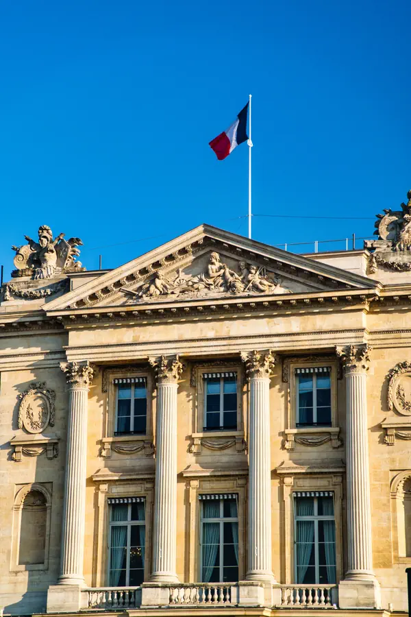 Façade néoclassique d'un bâtiment officiel parisien avec ses colonnes corinthiennes, son fronton triangulaire orné de bas-reliefs, et le drapeau français flottant au sommet.
