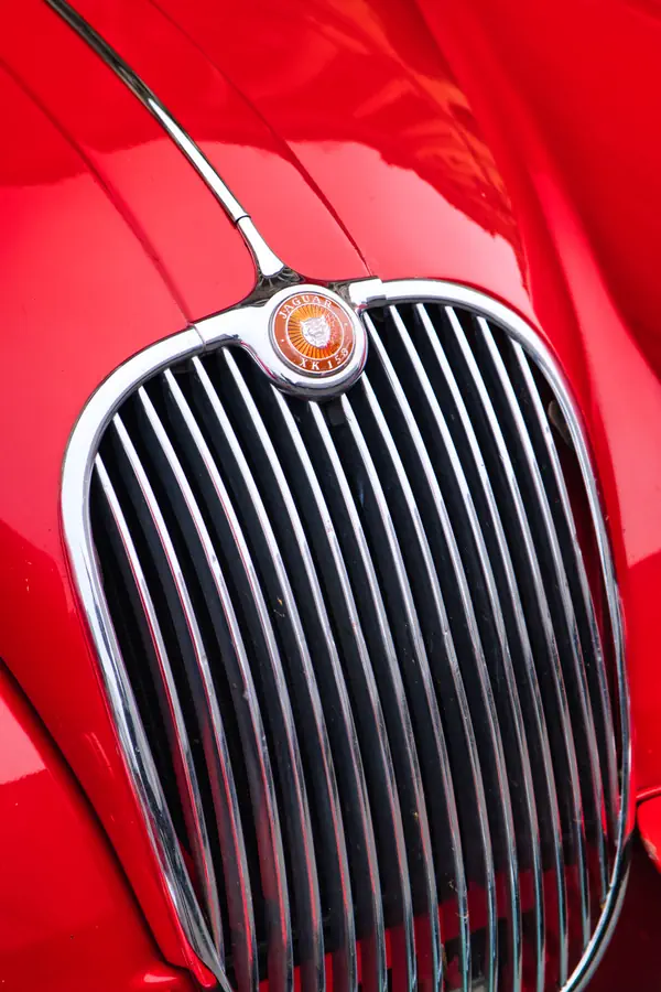 Calandre chromée d'une Jaguar classique avec ses barres verticales caractéristiques et son emblème rond orange au centre, sur une carrosserie rouge vif.