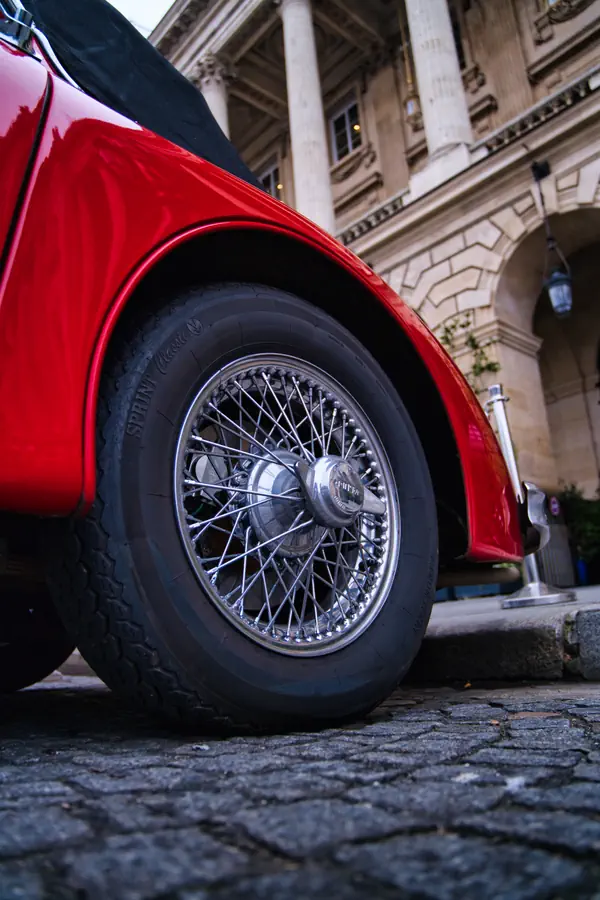 Détail de la roue à rayons chromés de la Jaguar XK150 rouge. La roue se détache sur l'aile rouge brillante et les pavés parisiens, avec les colonnes d'un bâtiment historique en arrière-plan.