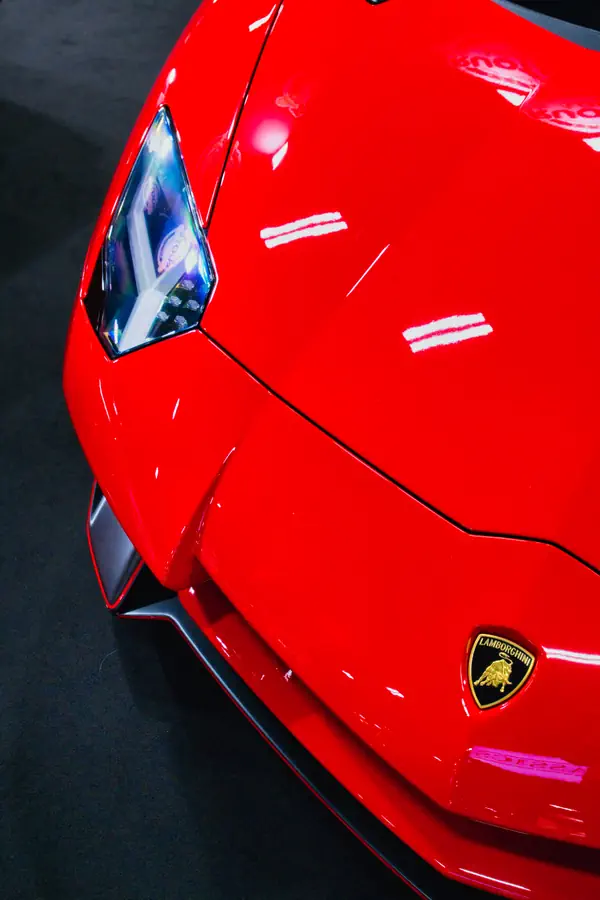Détail de l'avant d'une Lamborghini rouge montrant son phare angulaire caractéristique, les lignes agressives de son capot et l'emblématique écusson Lamborghini avec le taureau sur fond noir et or.