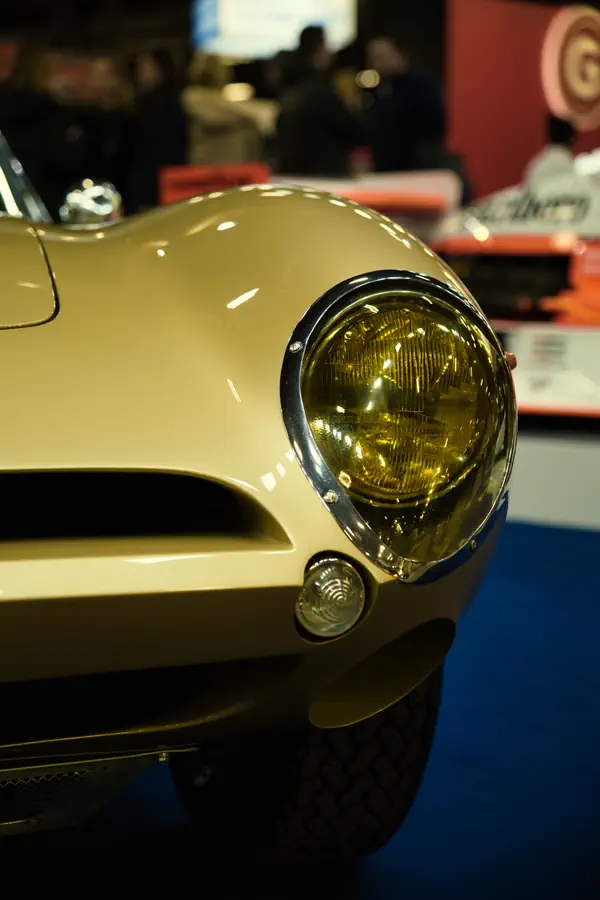 Détail de l'avant d'une voiture classique beige avec son phare rond chromé et son verre jaune caractéristique des voitures anciennes françaises, accompagné d'un petit feu de position.