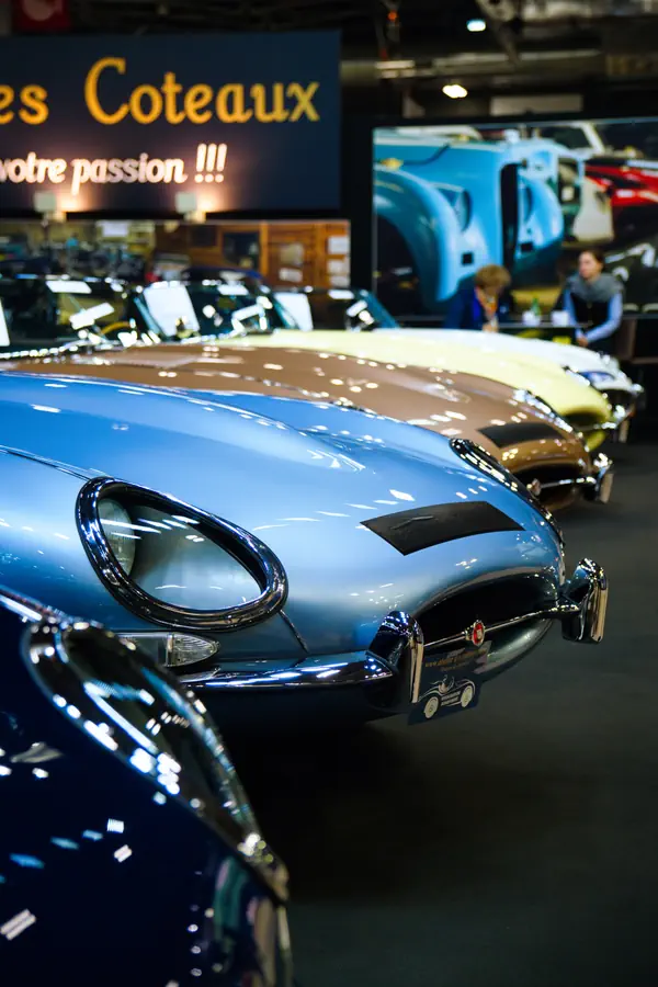 Avant d'une Jaguar Type E bleue exposée dans un salon automobile. D'autres voitures de collection sont alignées en arrière-plan, créant une belle exposition de voitures classiques.