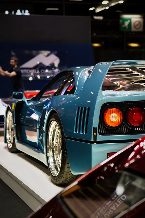 Arrière d'une Ferrari F40 bleu clair exposée dans un salon automobile, montrant ses feux arrière ronds caractéristiques, ses prises d'air latérales et sa jante en alliage chromée.