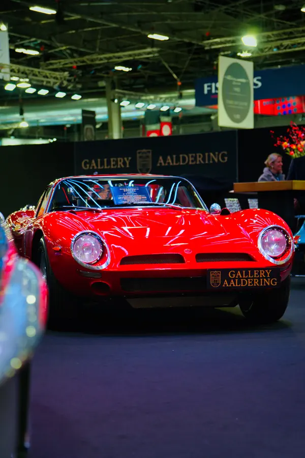 Bizzarrini 5300 GT rouge exposée sur le stand Gallery Aaldering. Cette voiture de sport italienne des années 60 se distingue par ses phares ronds caractéristiques et sa carrosserie basse et élancée.