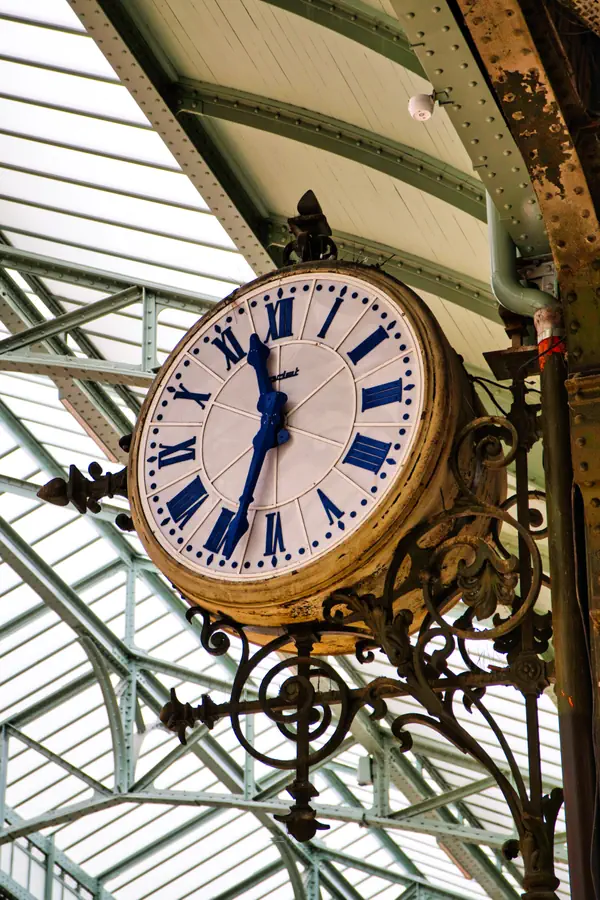 Horloge ancienne de gare avec son cadran blanc à chiffres romains bleus, montée sur un support en fer forgé décoratif.