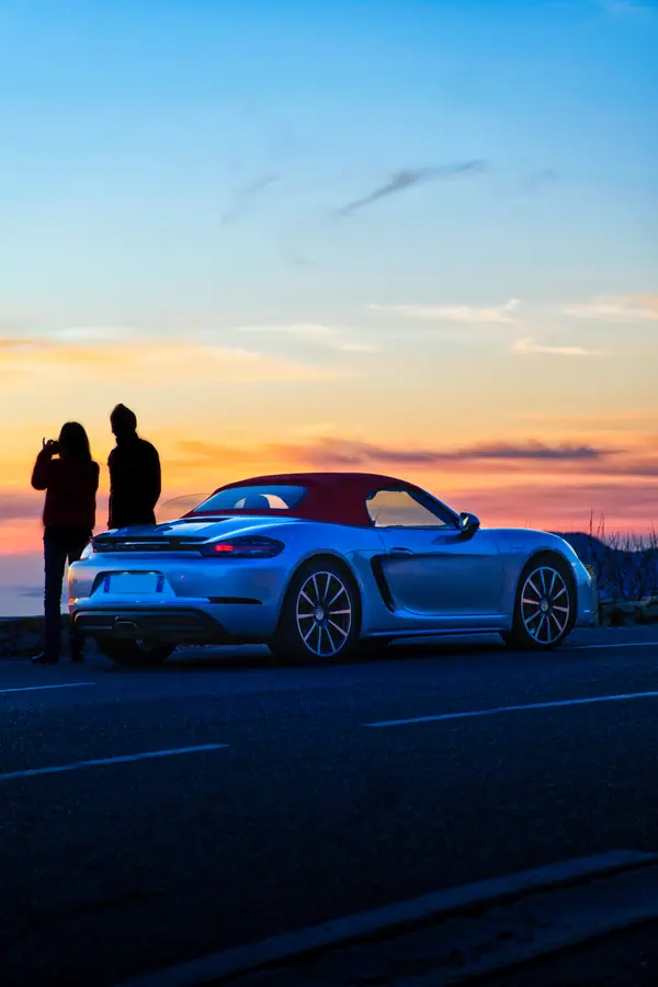 Porsche 718 Boxster argentée photographiée au coucher du soleil, capote rouge, vue de trois-quarts arrière, avec deux silhouettes admirant l'horizon orangé en arrière-plan.