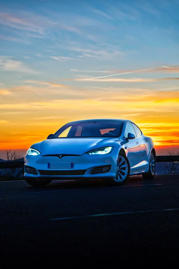 Tesla Model S blanche photographiée de trois-quarts avant au coucher du soleil, phares LED allumés, reflets orangés du ciel sur la carrosserie.