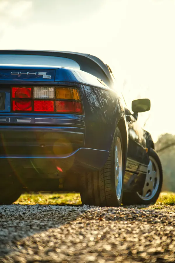 Détail arrière d'une Porsche 944 S2 bleue, photographiée en contre-jour au coucher du soleil, mettant en valeur les feux arrière caractéristiques et la jante en alliage, avec des reflets dorés sur le sol.
