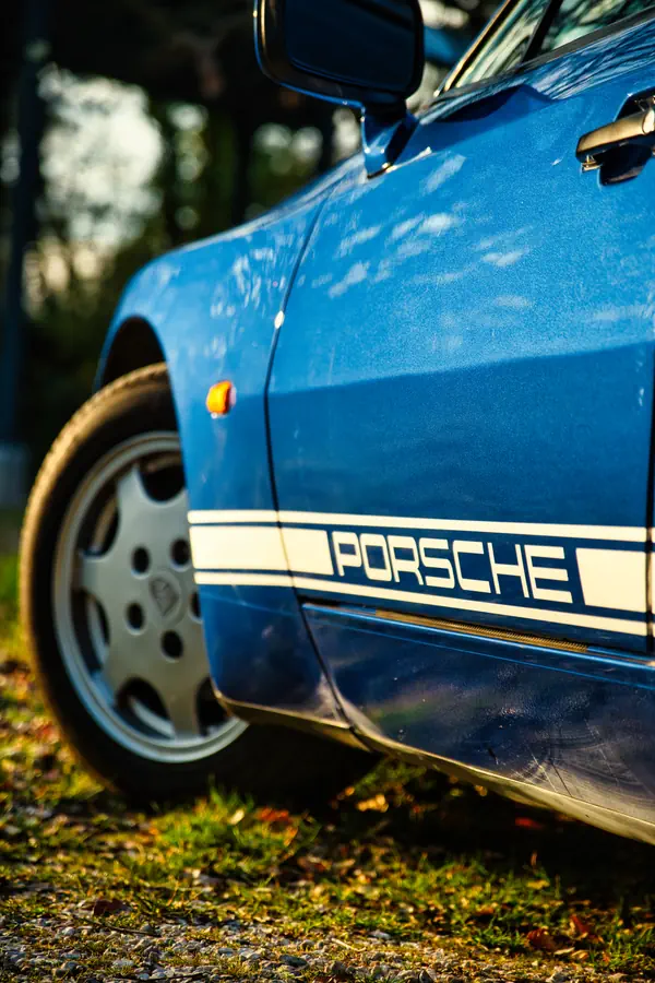 Détail de la portière d'une Porsche 944 bleue montrant le bandeau latéral décoratif blanc avec l'inscription "PORSCHE", répétiteur de clignotant orange et jante en alliage caractéristique visible en arrière-plan.