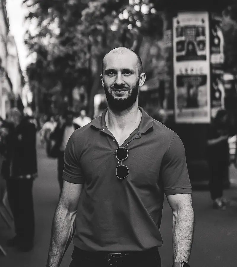 Portrait en noir et blanc d'un homme souriant dans une rue, portant un polo et des lunettes de soleil accrochées au col.